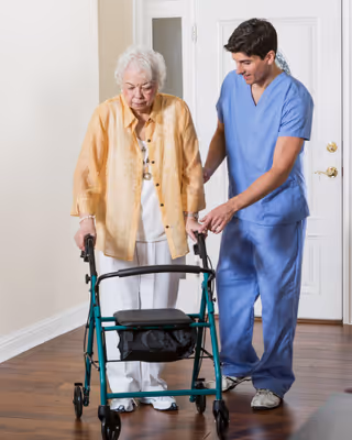 An elderly woman using a walker is being assisted by a male caregiver dressed in blue scrubs inside a residential setting with wooden floors and a white door in the background.