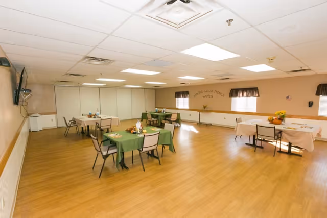 A spacious dining room with several tables covered with green and beige tablecloths, each set with chairs and small centerpieces. The room has light wood flooring, beige walls, and windows with dark valances. A wall-mounted TV is visible on the left side, and the far wall has the phrase 'Making Great Things Happen' displayed above the windows.