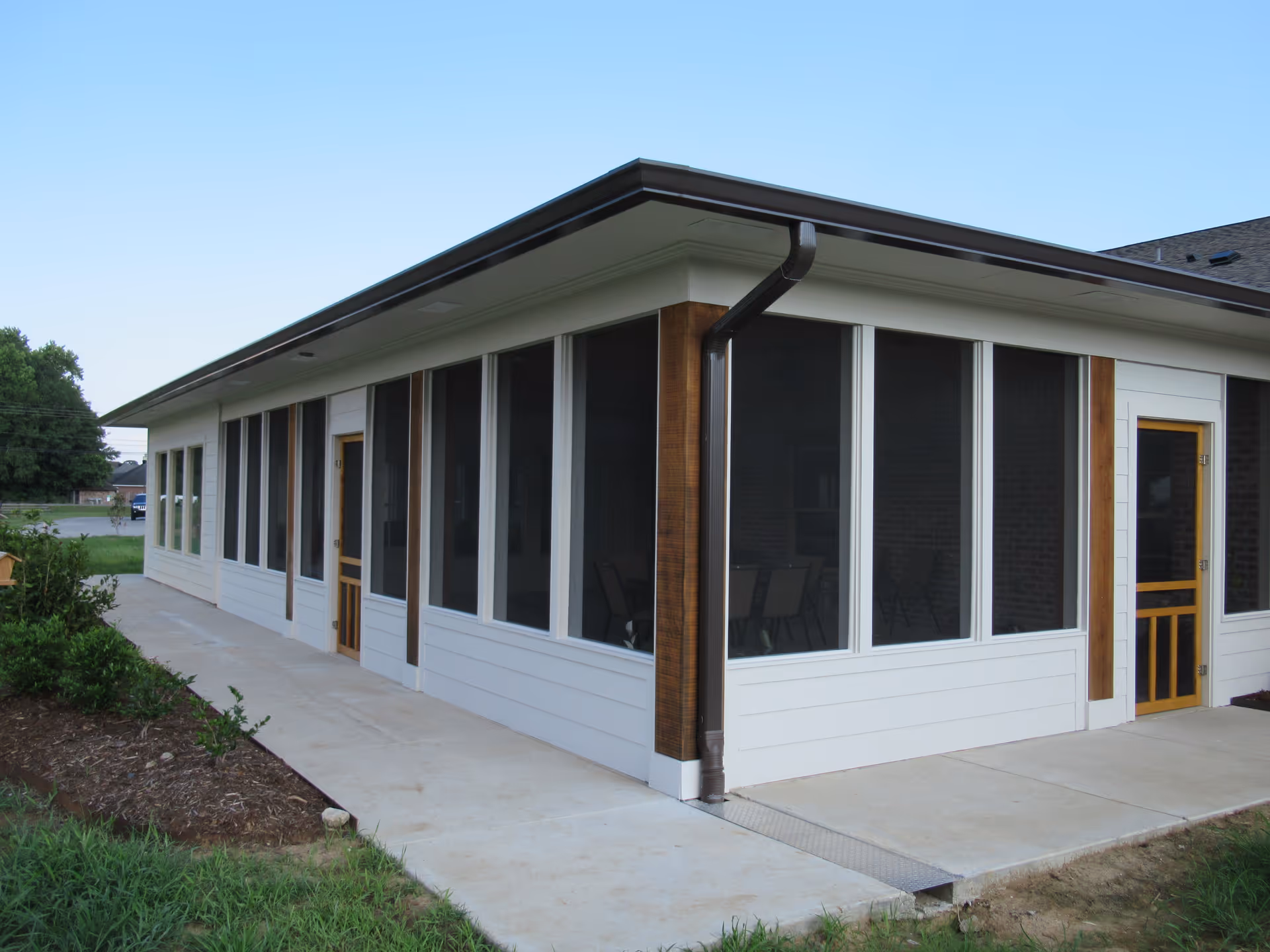 Exterior view of a single-story building with large screened windows and two wooden doors, surrounded by a concrete walkway and some greenery under a clear blue sky.