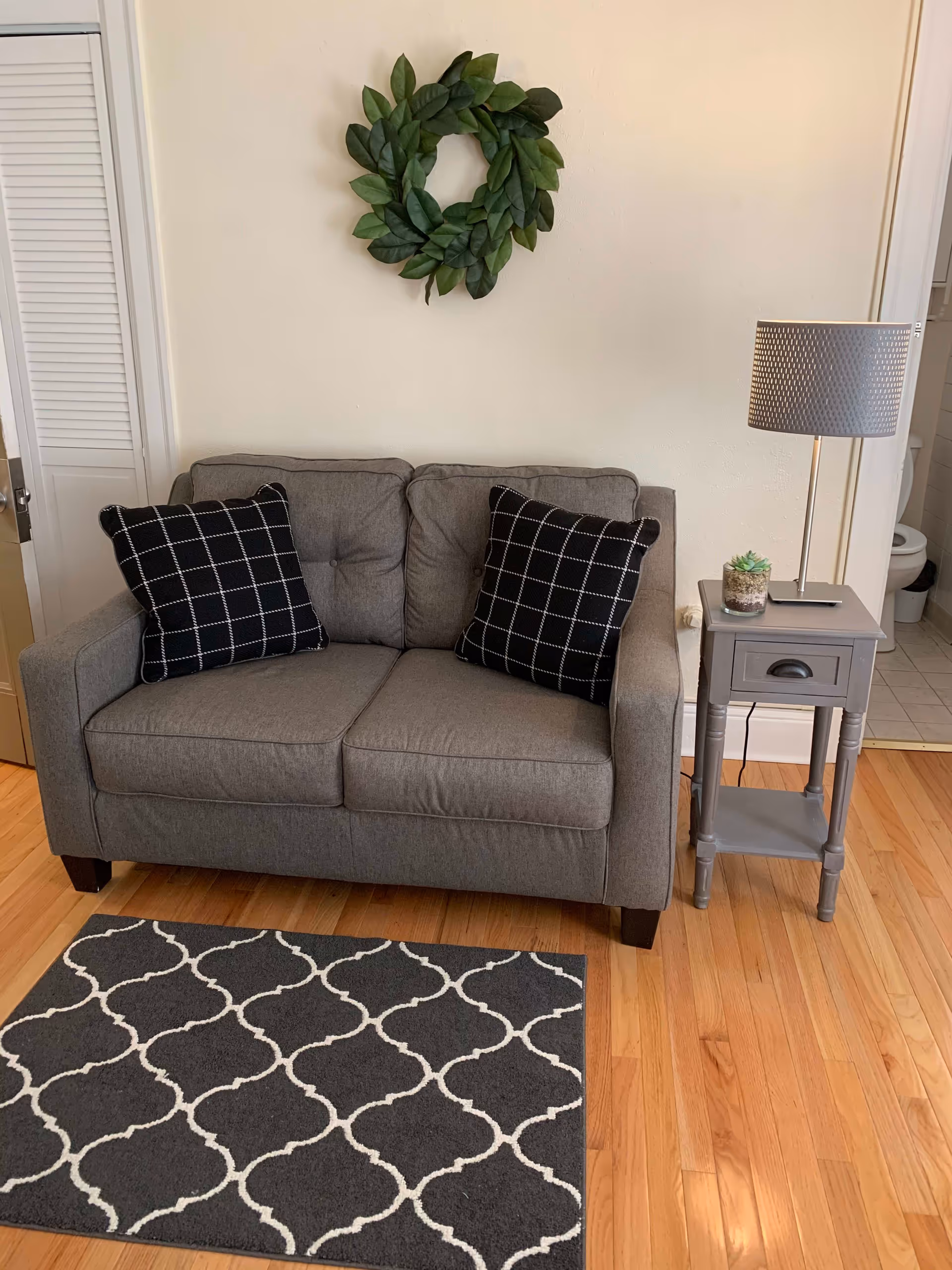 A small gray loveseat with two black and white checkered pillows is placed against a beige wall. Above the loveseat hangs a decorative green wreath. To the right of the loveseat is a gray side table with a drawer, holding a small potted succulent and a gray table lamp. The floor is wooden with a small dark gray rug featuring a white geometric pattern in front of the loveseat. A bathroom with a visible toilet is partially seen through an open door on the right.