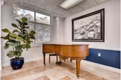 A room with a wooden grand piano placed on a tiled floor. There is a large window with blinds partially open, allowing natural light to enter. A large green potted plant is positioned near the window. On the wall, there is a framed black and white photograph depicting a street scene with cars and people. The lower half of the wall is painted dark blue with a white chair rail molding.