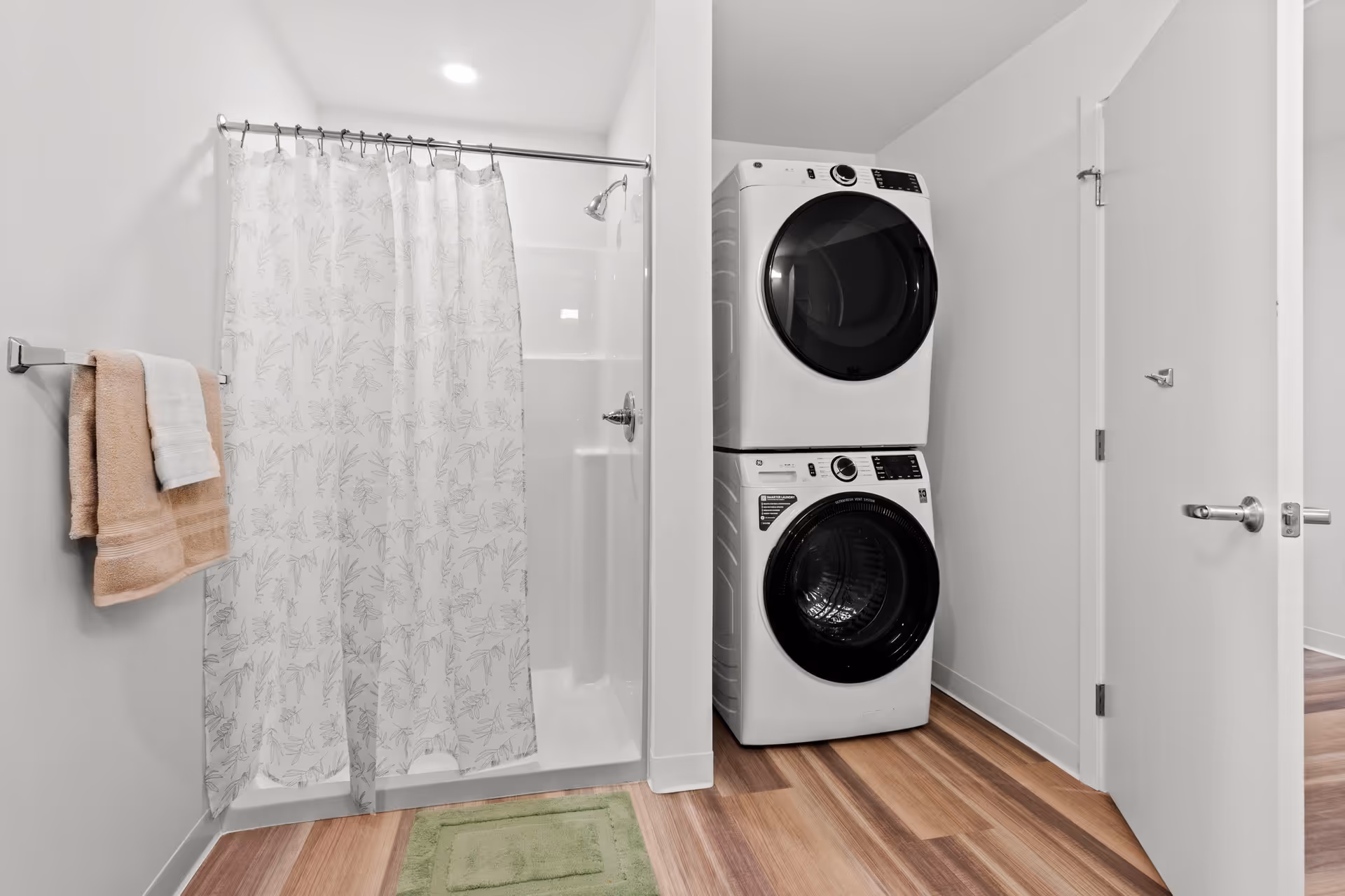 A bathroom with a shower and patterned curtain, towel rack, stacked washer and dryer, and wood-look flooring.