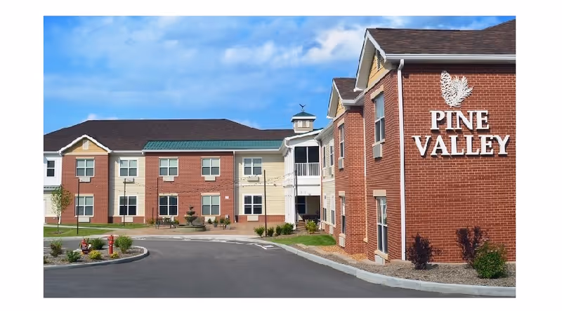 Exterior view of Pine Valley Assisted Living facility showing a two-story brick and siding building with multiple windows, a driveway, and landscaped areas with small trees and shrubs under a partly cloudy blue sky.