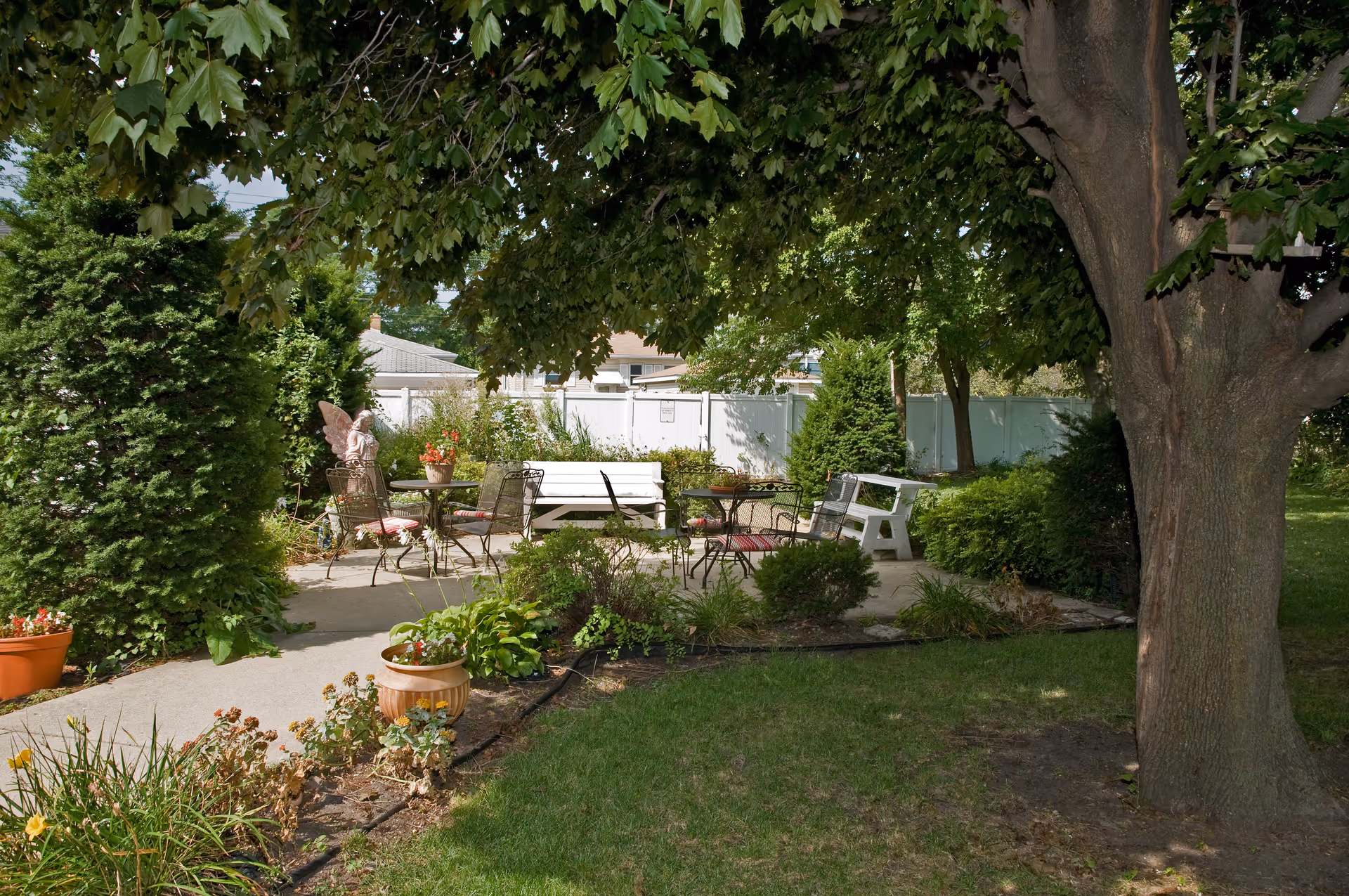Shaded outdoor patio garden with metal chairs, tables, benches and potted plants under a large tree.