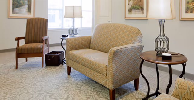 A cozy sitting area with a patterned loveseat, a striped armchair, a round wooden side table with a decorative lamp, and framed artwork on the walls. The room is well-lit with natural light coming through the windows.