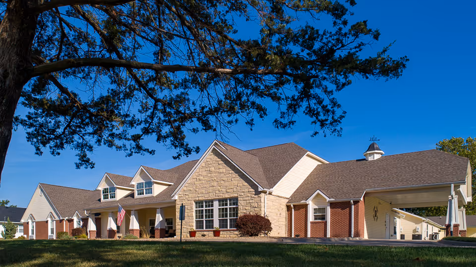 Exterior view of a single-story assisted living facility building with a combination of brick and stone facade, multiple windows, a covered entrance, and a large tree in the foreground under a clear blue sky.