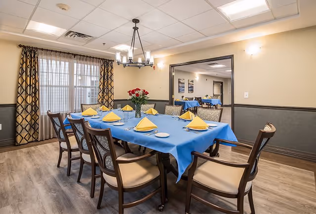 A dining room with a rectangular table covered with a blue tablecloth, set with yellow folded napkins, plates, and silverware. There are eight wooden chairs with cushioned seats around the table. A vase with red flowers is placed in the center. The room has light-colored walls with a dark wainscot, wood flooring, patterned curtains on a window, and a chandelier hanging above the table. Another dining area with similar tables and chairs is visible through an open doorway.