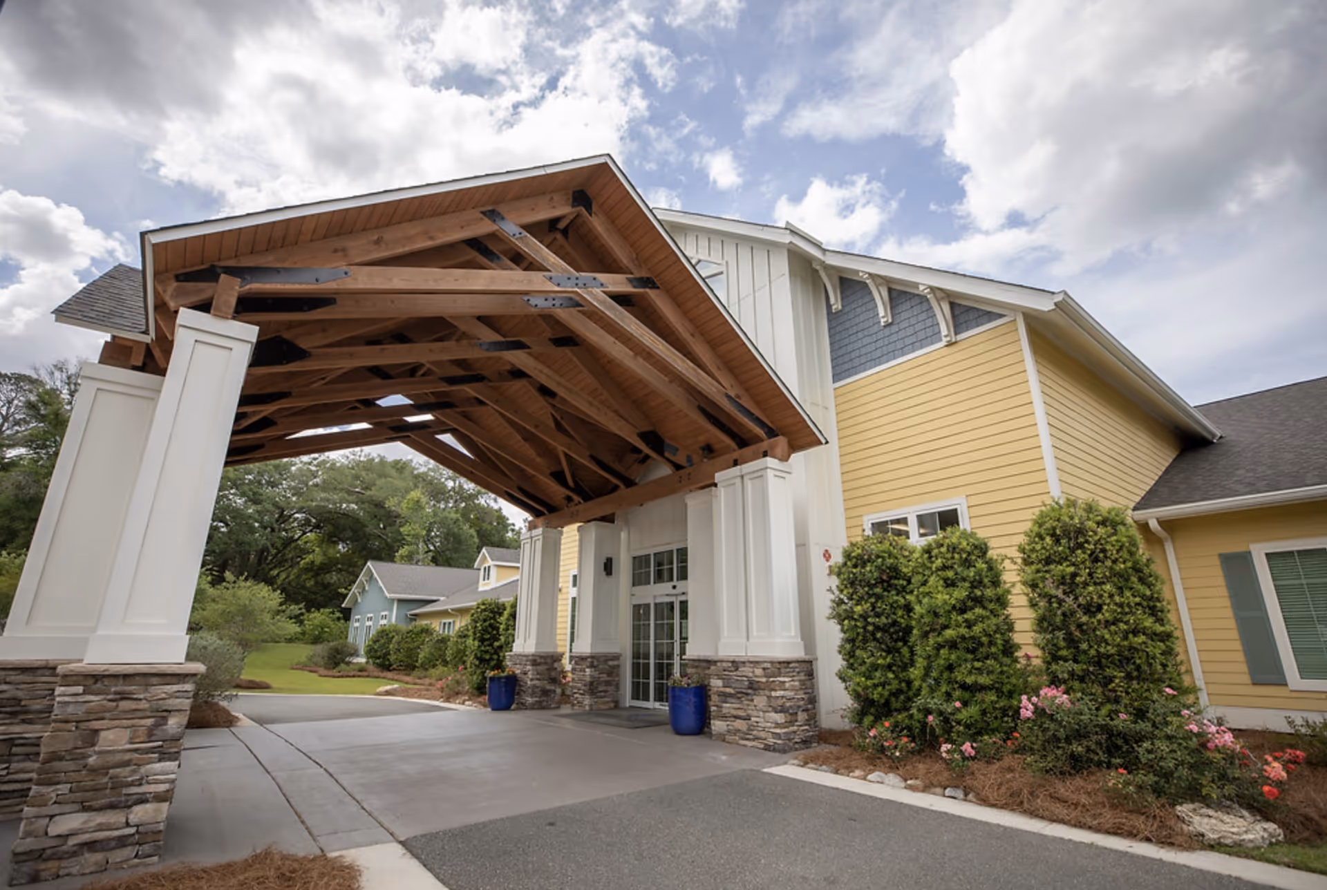 Entrance of a senior living facility with a large wooden covered driveway supported by white columns with stone bases. The building exterior is painted yellow with white trim and has neatly trimmed bushes and flowers along the side. The sky is partly cloudy.