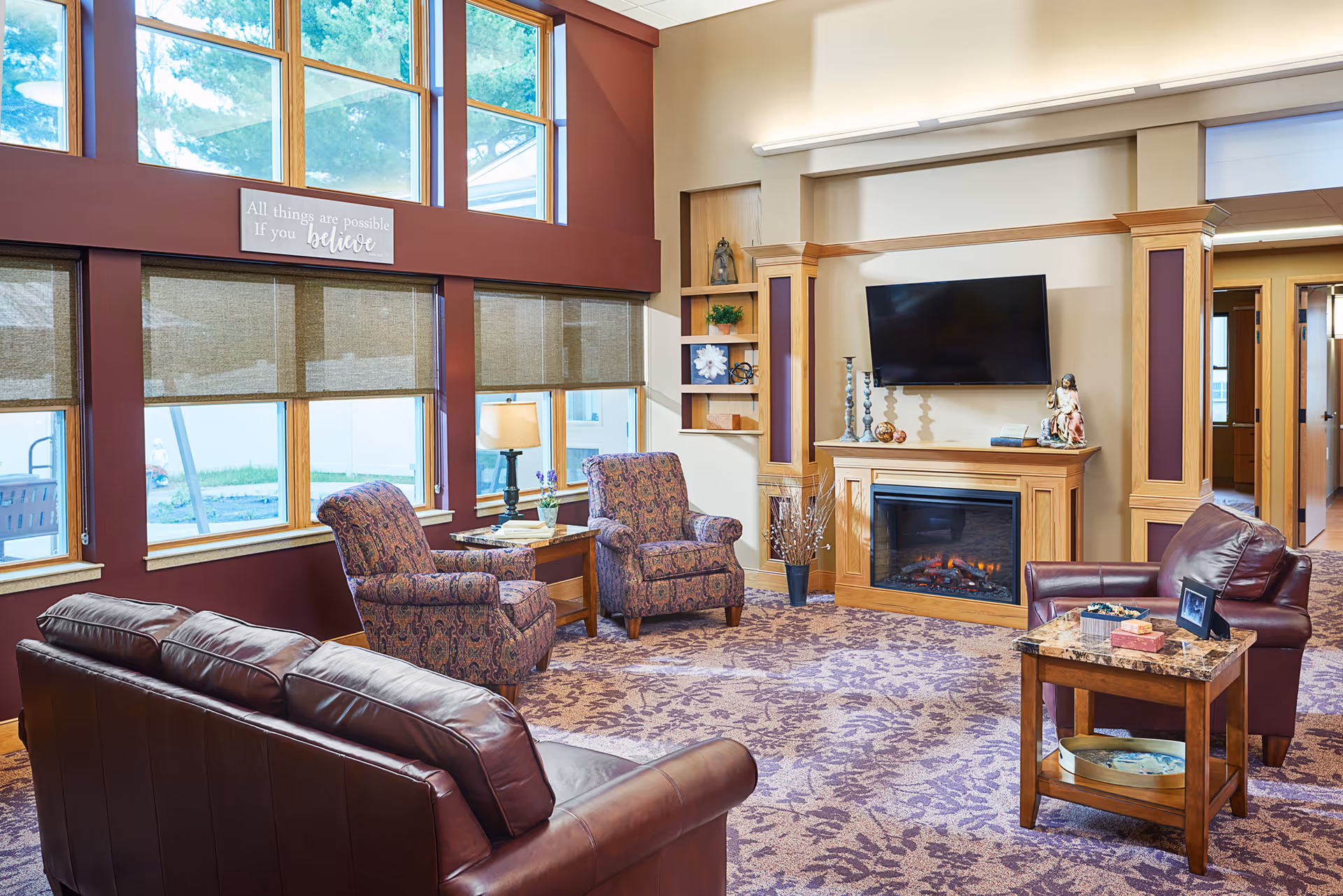 Cozy senior living lounge with leather sofas, patterned chairs, a fireplace, and large windows.