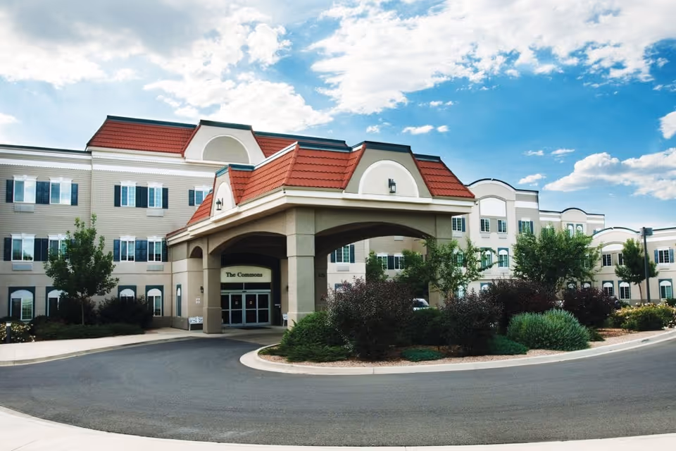 Exterior view of The Commons of Hilltop senior living facility showing a large building with beige walls, red roof accents, multiple windows, and a covered entrance with landscaping and a circular driveway under a partly cloudy blue sky.