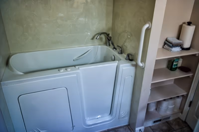 Walk-in bathtub with a side door, grab bar, faucet, and nearby shelving holding paper towels and toilet paper.