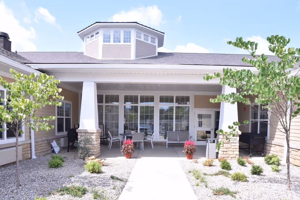 Covered entrance of a senior living building with a porch, chairs, potted plants, and landscaped gravel beds.