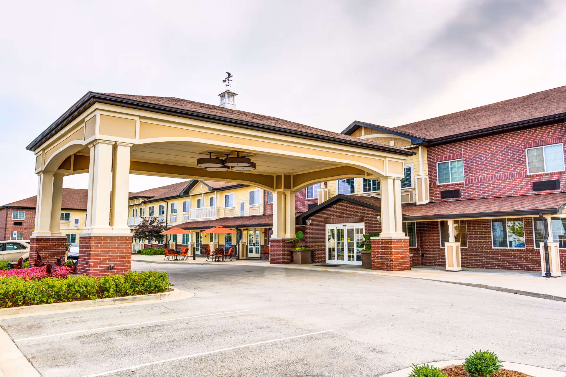 Covered porte-cochère entrance of a multi-story senior living building with brick and yellow siding, outdoor seating, and landscaping.