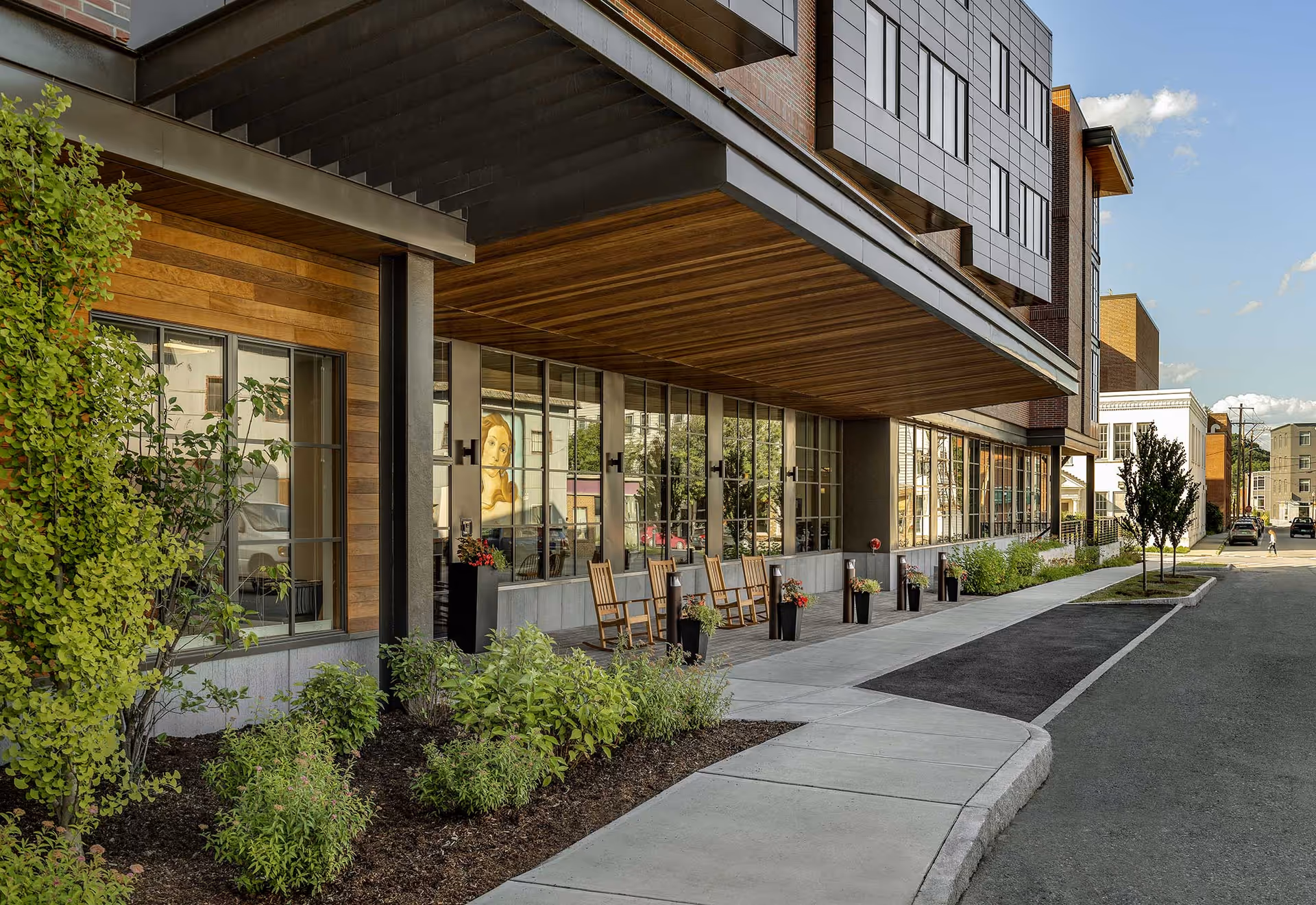 Exterior view of The Village at White River Junction building with a covered porch area featuring wooden rocking chairs and potted plants. The building has large windows reflecting the street and nearby buildings, with a sidewalk and landscaped greenery in front.