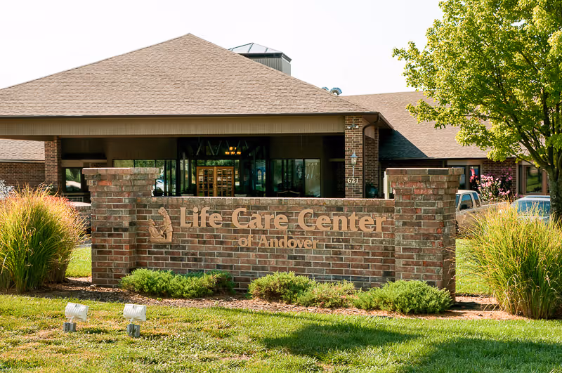 Brick sign reading 'Life Care Center of Andover' on a lawn in front of the facility entrance.