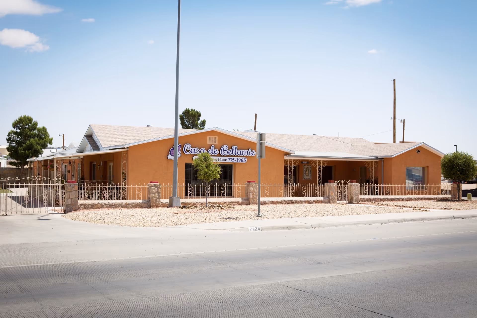Single-story orange assisted living building 'Casa de Bellamie' with a front fence, desert landscaping, and a street in the foreground.