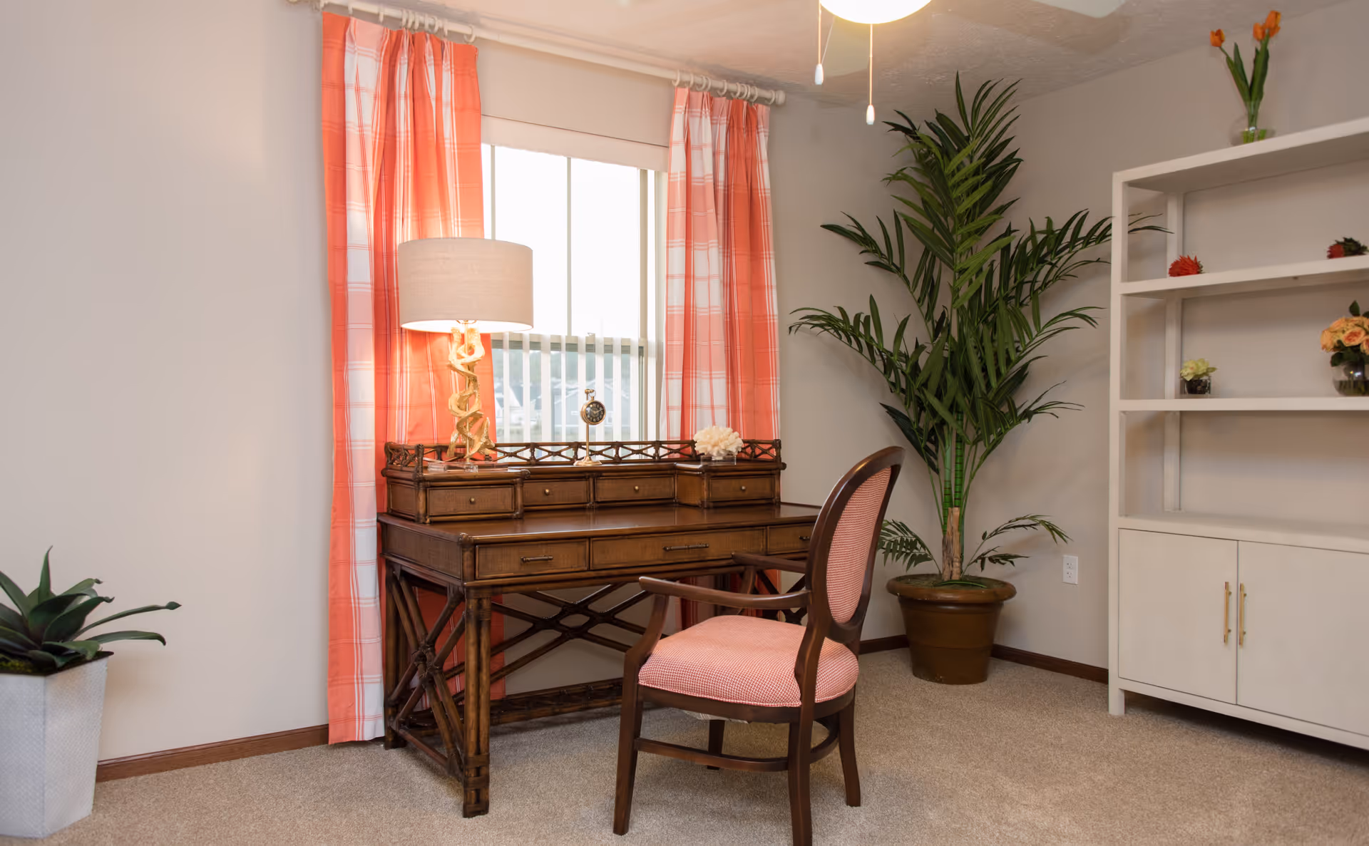 A cozy interior room featuring a wooden desk with drawers, a table lamp with a beige shade, and a small clock on the desk. The desk is positioned in front of a window with coral and white plaid curtains. A wooden chair with coral upholstery is placed in front of the desk. To the right, there is a tall potted plant and a white shelving unit with decorative flowers. Another smaller potted plant is on the floor to the left.