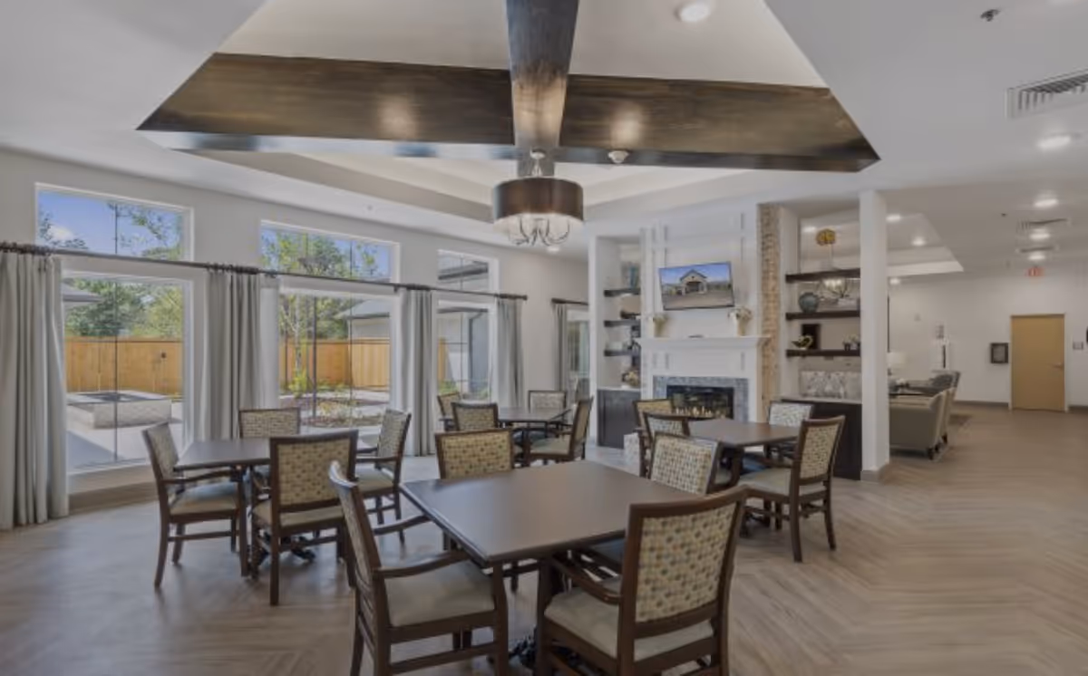 A bright and spacious common area with several square tables and cushioned chairs arranged around them. Large windows with curtains allow natural light to fill the room and provide a view of an outdoor patio area with a wooden fence. The ceiling features a decorative wooden beam and a chandelier. There is a white fireplace with a mounted TV above it and built-in shelves with decorative items on the right side. The flooring is light wood, and the overall decor is modern and welcoming.