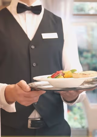 A waiter dressed in a white shirt, black vest, and black bow tie holding a silver tray with a plate of food that includes a salad and a piece of bread, with a blurred background of a dining area.