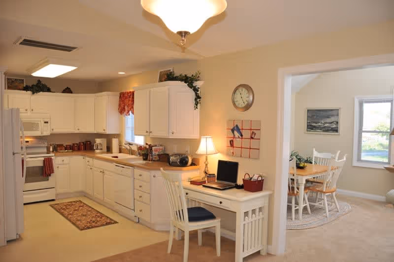 A bright kitchen with white cabinets, white appliances including a refrigerator, stove, microwave, and dishwasher. There is a small desk area with a chair, lamp, and laptop on it. The kitchen opens into a dining area with a wooden table and white chairs, a window, and a painting on the wall.