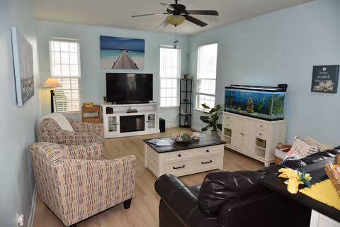Bright furnished living room with a TV on a white console, coffee table, armchairs, a leather sofa, and a large aquarium.