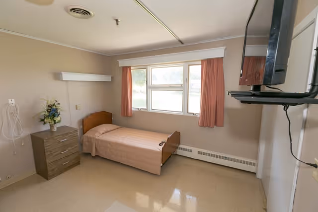 A simple, clean nursing home bedroom with a single bed covered in a light brown bedspread, a small wooden nightstand with a vase of flowers, a window with peach-colored curtains, and a wall-mounted television.