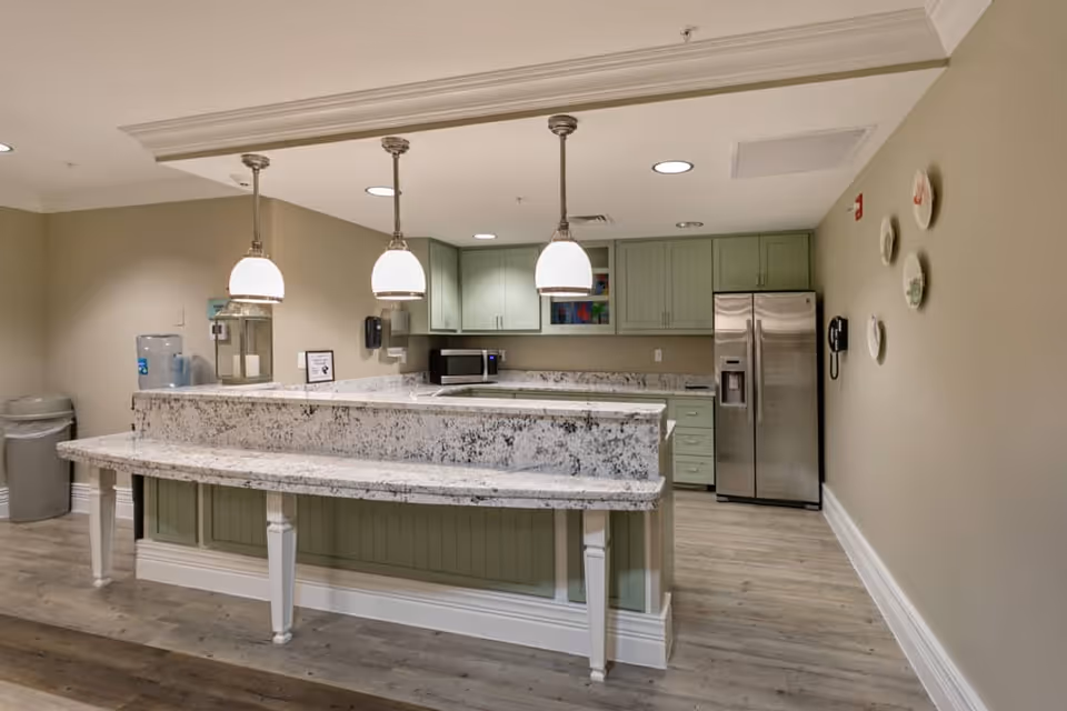 Interior view of a kitchen area in an assisted living facility featuring green cabinets, a stainless steel refrigerator, a microwave, a granite countertop with a breakfast bar, three hanging pendant lights, and decorative plates on the wall.