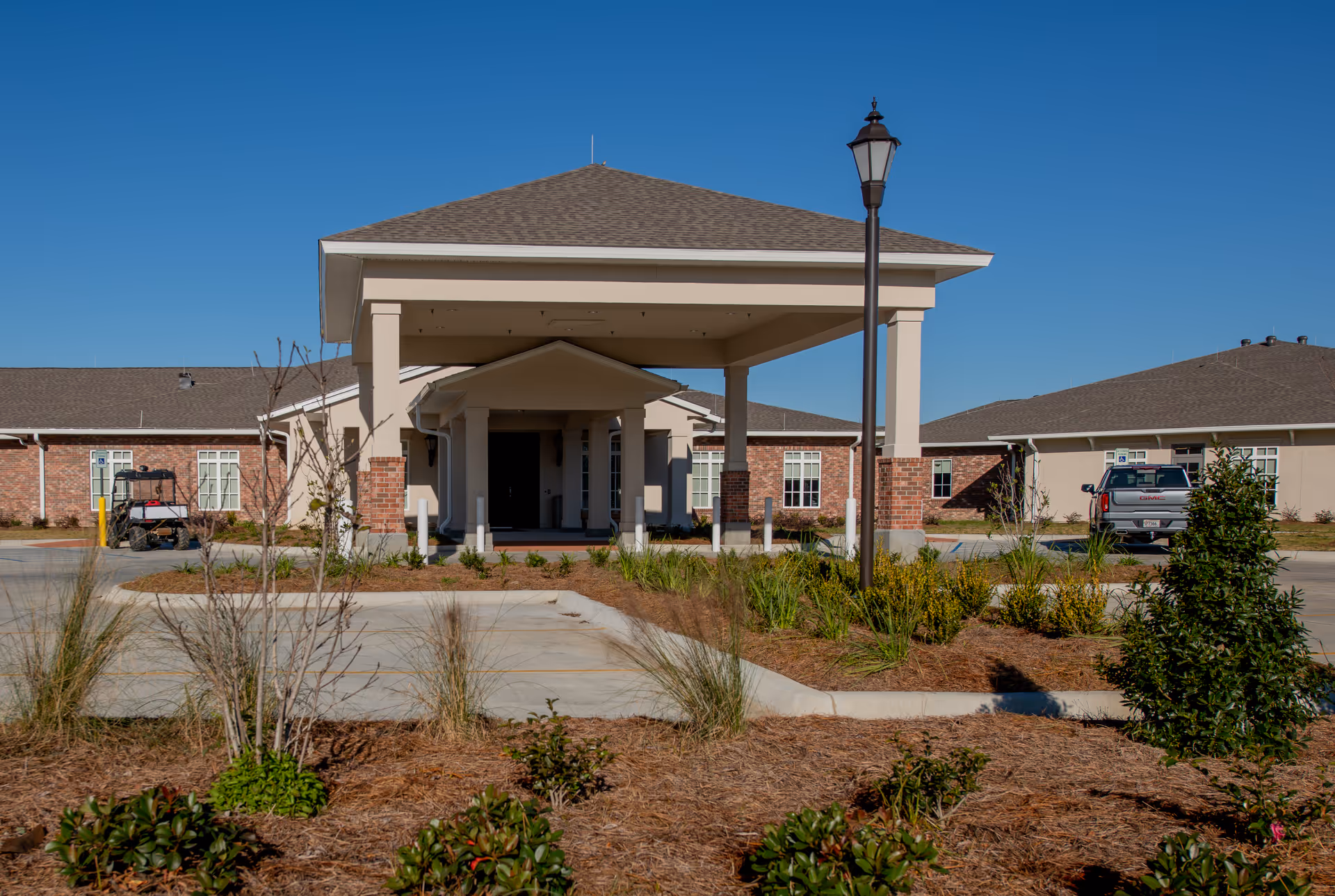 Front exterior view of Thibodaux Healthcare and Rehabilitation Center showing a covered entrance with columns, a lamp post, landscaped garden beds, and a clear blue sky.