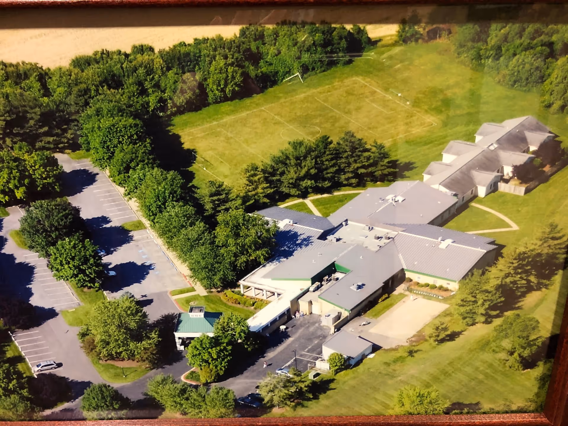 Aerial view of Regency Park Senior Living Community showing a large building complex surrounded by green lawns, trees, and a parking lot. There is a soccer field visible behind the building.