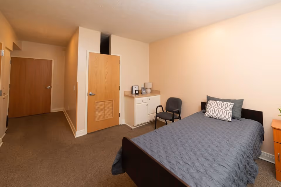 A simple bedroom in a senior living facility with a single bed covered in a gray quilt and two pillows, a black chair, a small white cabinet with framed photos and a lamp, beige walls, and two wooden doors.