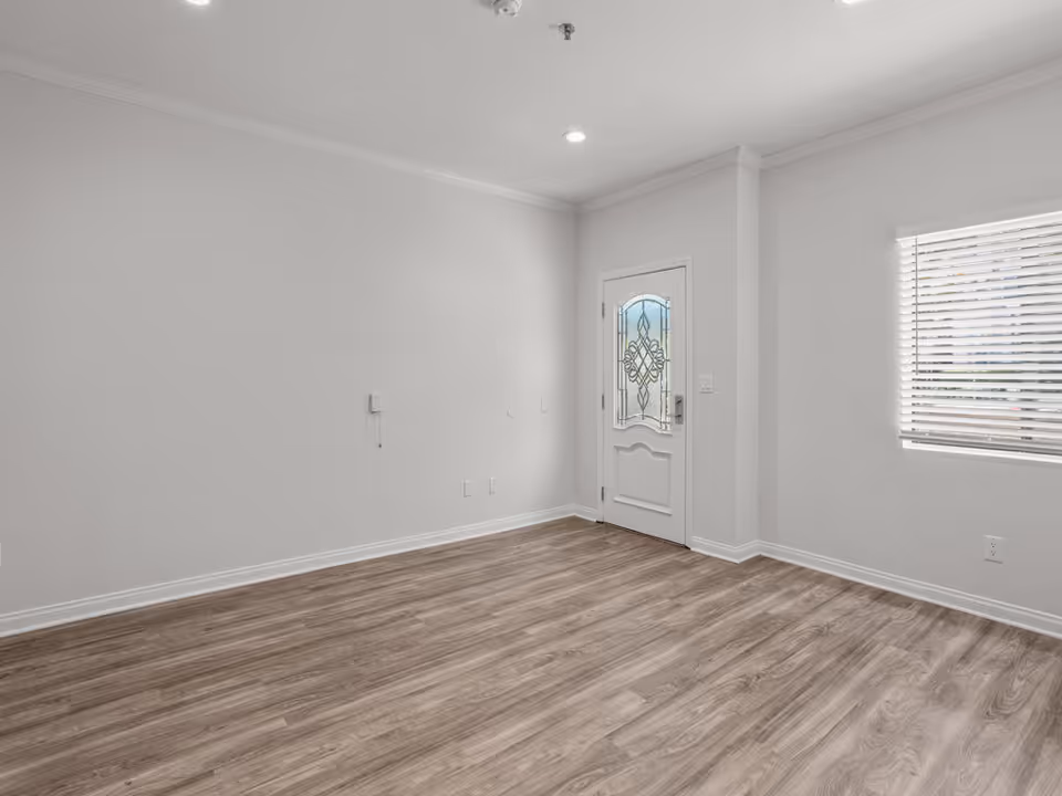 Empty room with light gray walls, wood-patterned flooring, a white door with decorative glass, and a window with white blinds allowing natural light to enter.