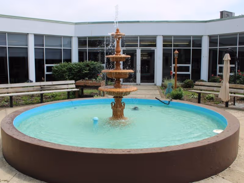 Outdoor courtyard area with a large round water fountain in the center, surrounded by benches, plants, and a building with large windows in the background.