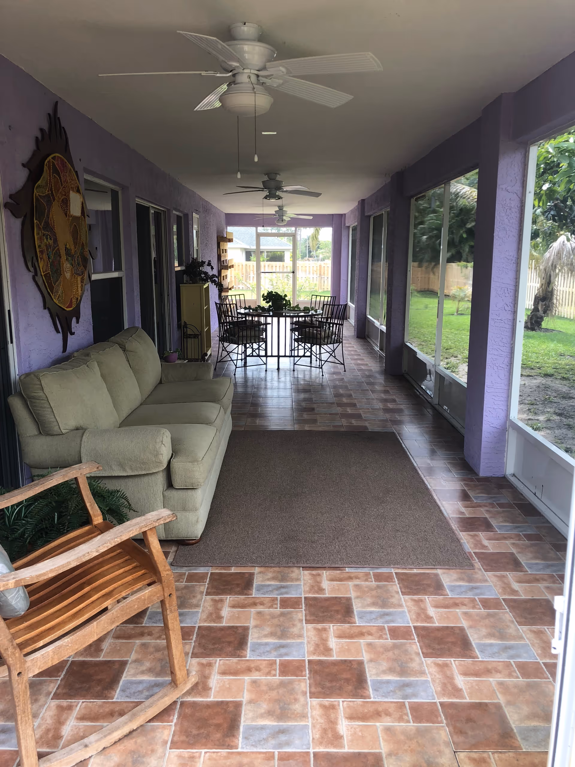 A long, covered patio area with purple walls and tiled floor. The space features a green couch, a wooden rocking chair, a brown rug, and a dining table with four metal chairs. Ceiling fans are mounted overhead, and large screened windows provide a view of a grassy yard with trees and a wooden fence.