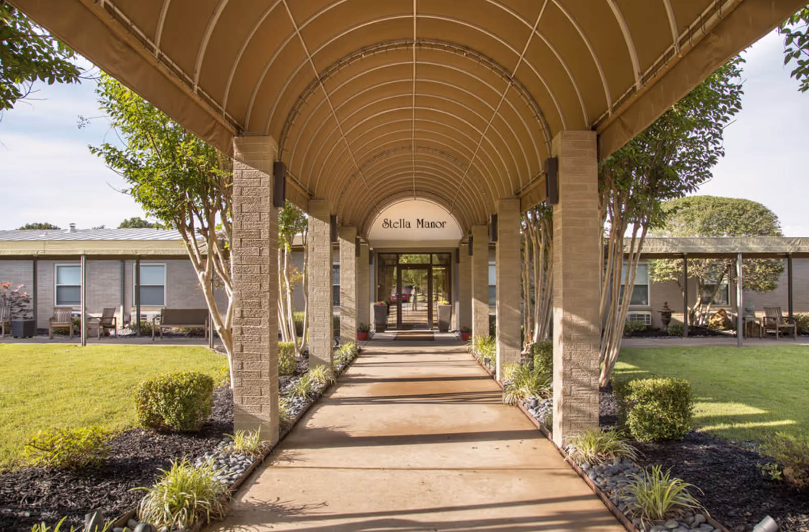 Covered walkway with arched roof leading to the entrance of Stella Manor, surrounded by neatly trimmed bushes, trees, and green lawn on either side.