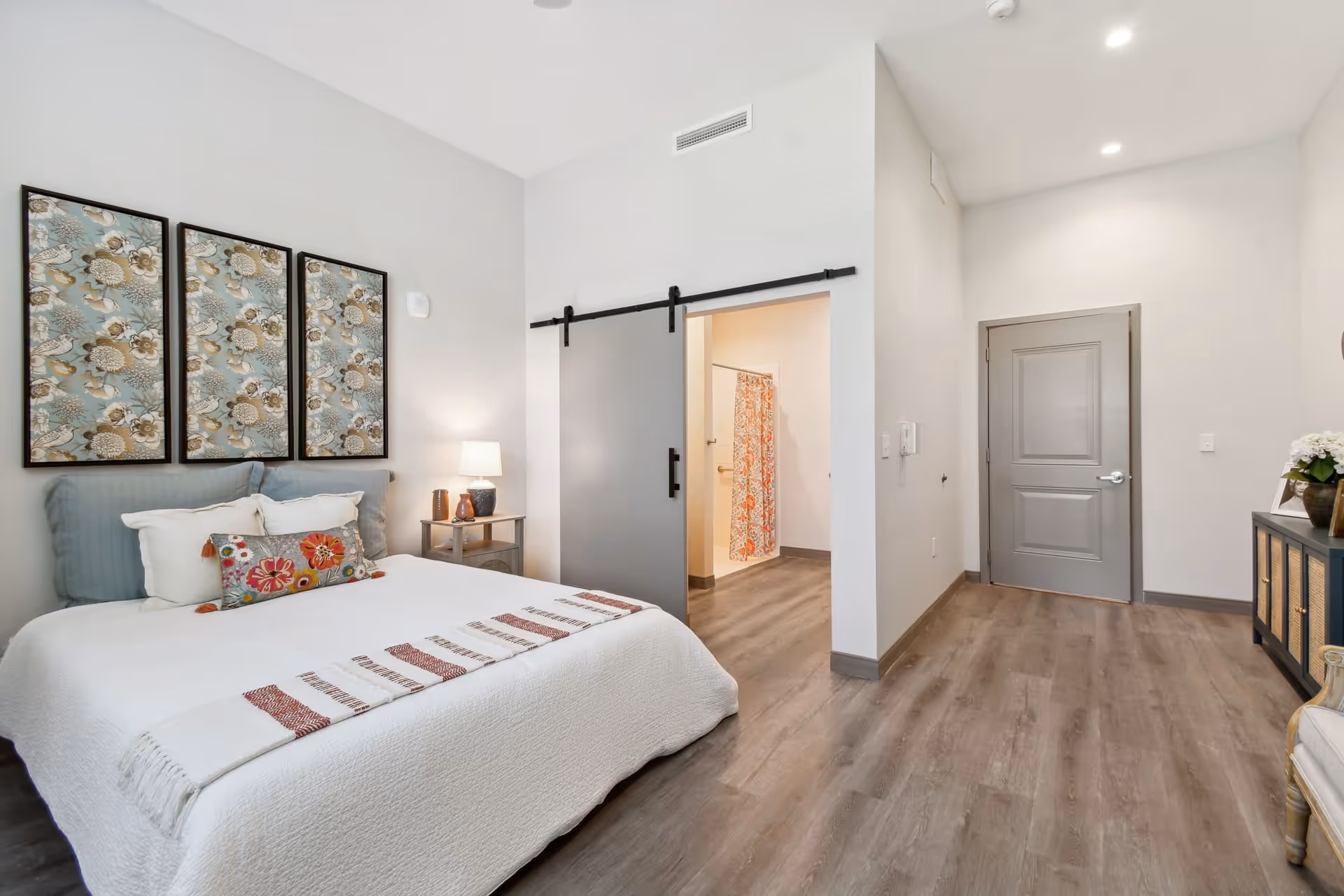 Bright bedroom with a made bed, three floral wall panels above the headboard, a sliding barn door to a bathroom, and gray wood-look flooring.