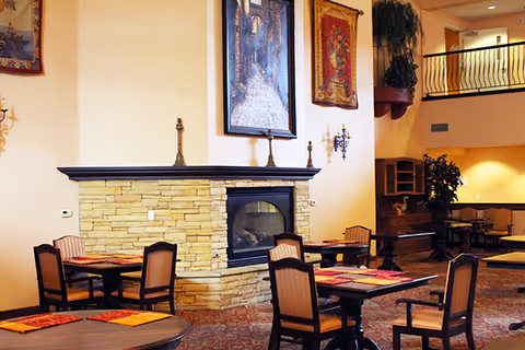 Interior view of a common dining area with multiple tables and chairs arranged around a stone fireplace. The walls are decorated with framed artwork and tapestries, and there is a balcony railing visible on the upper level. The space has warm lighting and a cozy atmosphere.