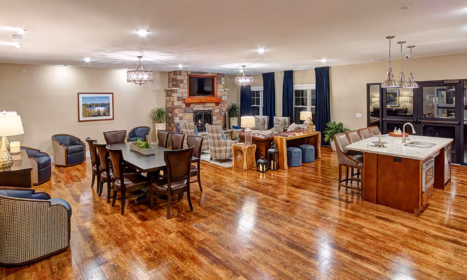 A spacious, well-lit common area featuring a dining table with chairs, a cozy seating area with armchairs and a stone fireplace with a mounted TV above it, and a kitchen island with bar stools and pendant lights. The room has wooden flooring and large windows with dark curtains.