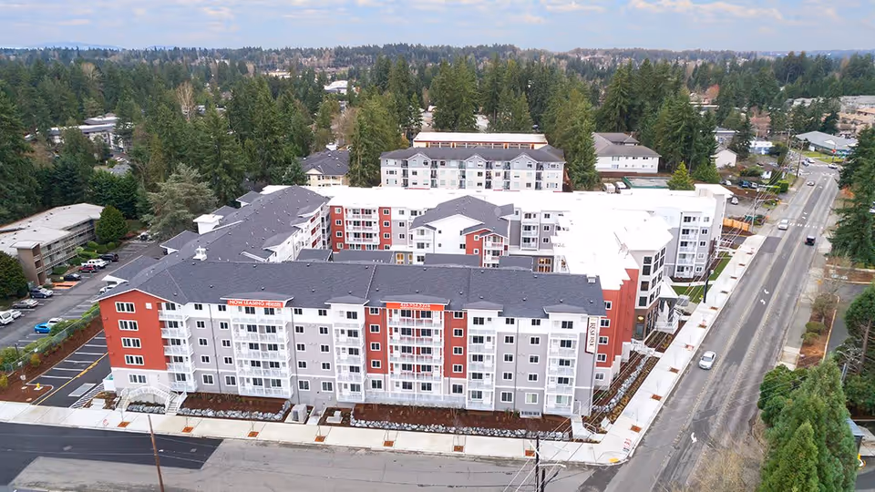 Aerial view of The Reserve At Lynnwood, a large multi-story residential building complex surrounded by trees and roads, with parking areas and sidewalks visible.