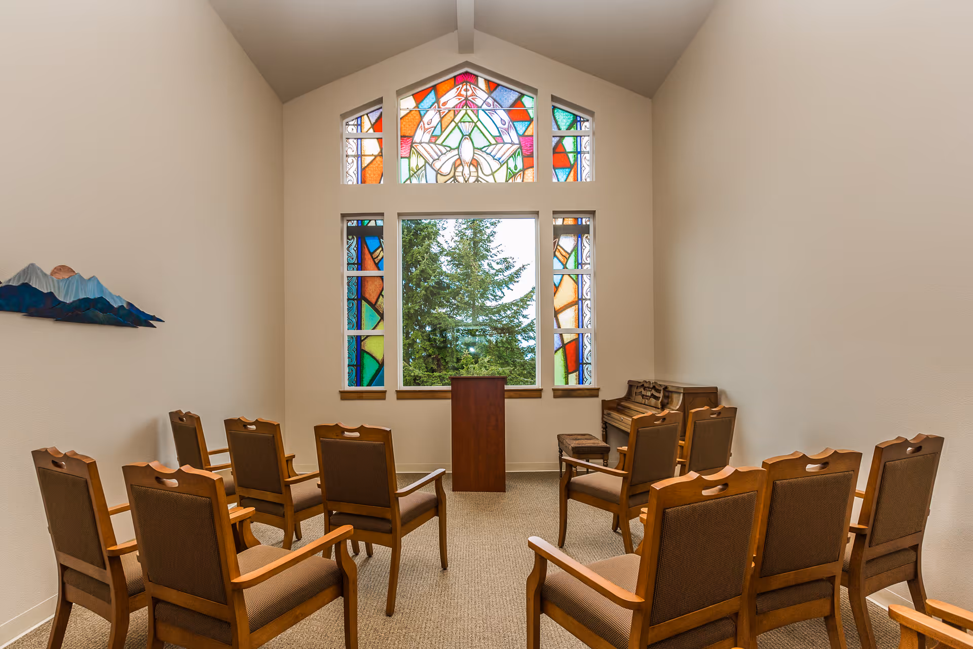 A small chapel or meditation room with wooden chairs arranged in rows facing a wooden podium. There is a piano with a stool to the right side of the room. The back wall features a large window with colorful stained glass panels surrounding a clear central window showing trees outside. The walls are light-colored and the room has a high ceiling.