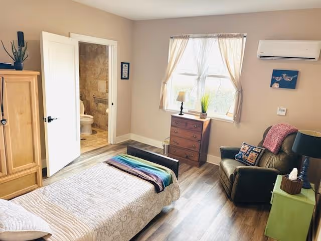 A cozy bedroom with a single bed covered in a beige quilt and a colorful blanket at the foot. To the left is a wooden wardrobe, and an open door reveals a bathroom with a toilet and tiled walls. A window with sheer curtains lets in natural light above a wooden chest of drawers with a lamp and a plant. To the right, there is a comfortable armchair with a pink throw and decorative pillows, next to a green side table holding a lamp and a tissue box. The room has wood flooring and beige walls.