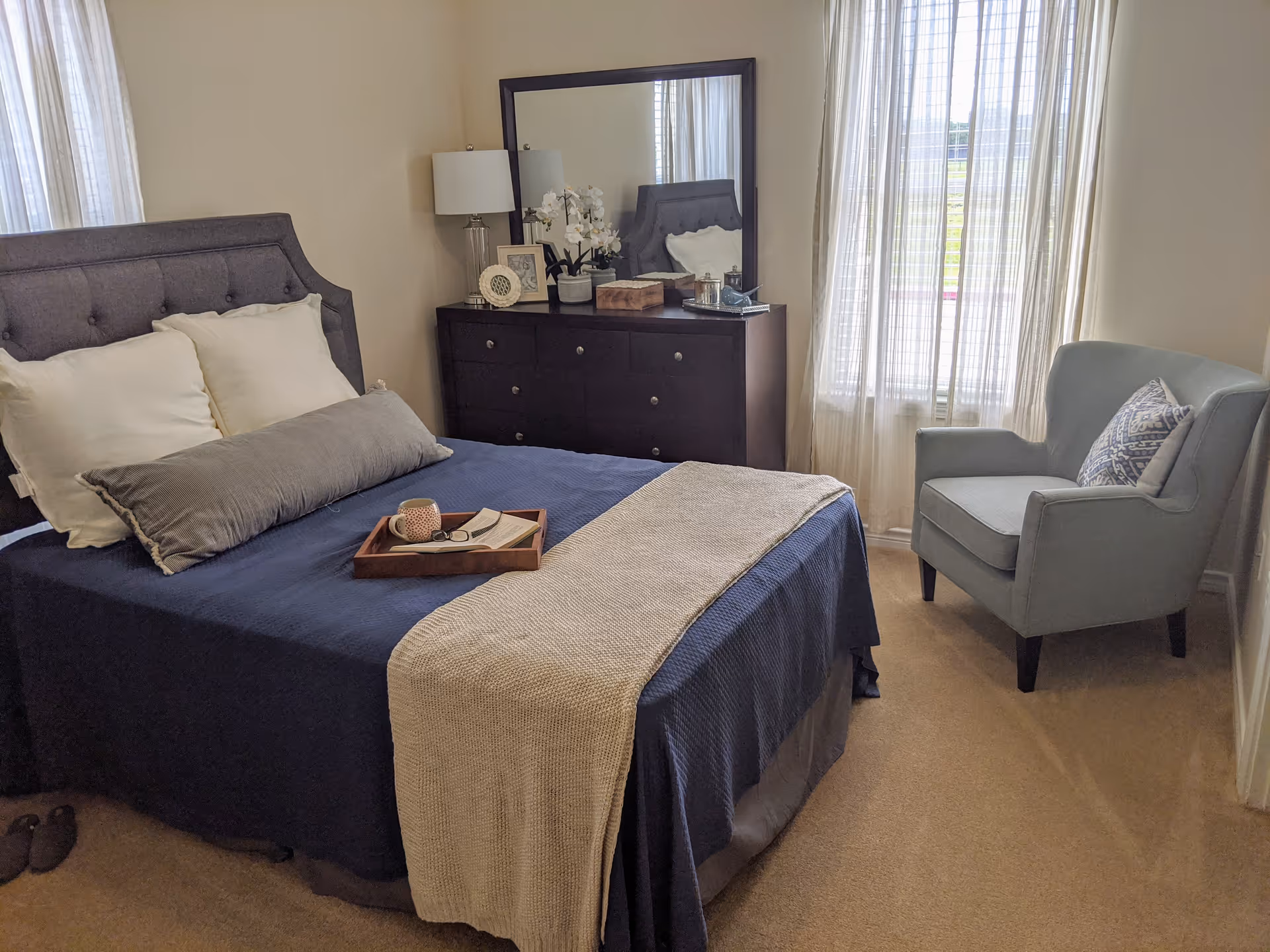 A cozy bedroom with a neatly made bed featuring a navy blue bedspread, white and gray pillows, and a beige throw blanket. A wooden tray with a cup, glasses, and an open book rests on the bed. To the left of the bed is a dark wooden dresser with a large mirror, a lamp, framed photos, and decorative items. A light blue armchair with a patterned pillow sits near a window with sheer white curtains, allowing natural light to fill the room.