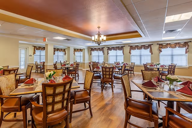 A spacious dining room with multiple wooden tables and chairs arranged neatly. Each table is set with red folded napkins, glasses, and small floral centerpieces. The room has large windows with valance curtains, a chandelier hanging from a recessed ceiling, and wood flooring.