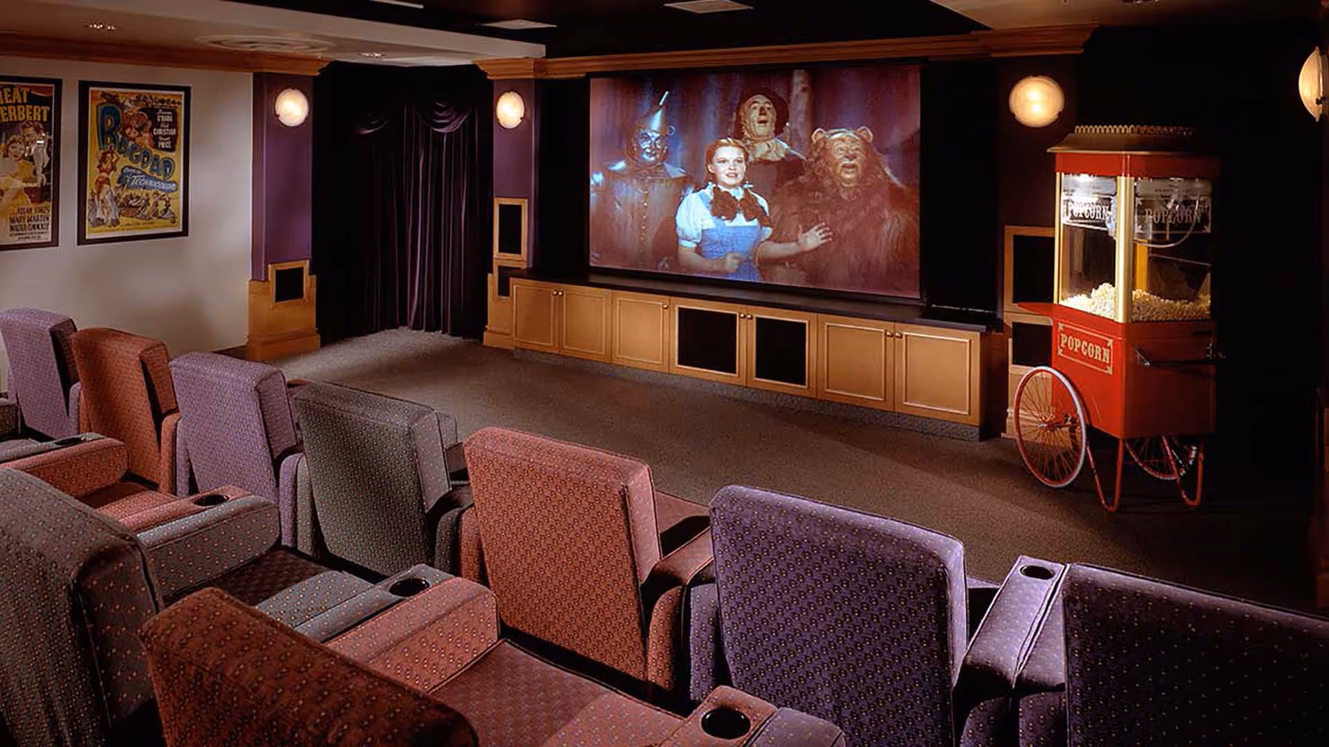 A cozy movie theater room with several rows of upholstered armchairs facing a large screen showing a scene from The Wizard of Oz. The room has dark walls with framed movie posters, wall sconces for lighting, and a vintage-style popcorn machine on the right side.