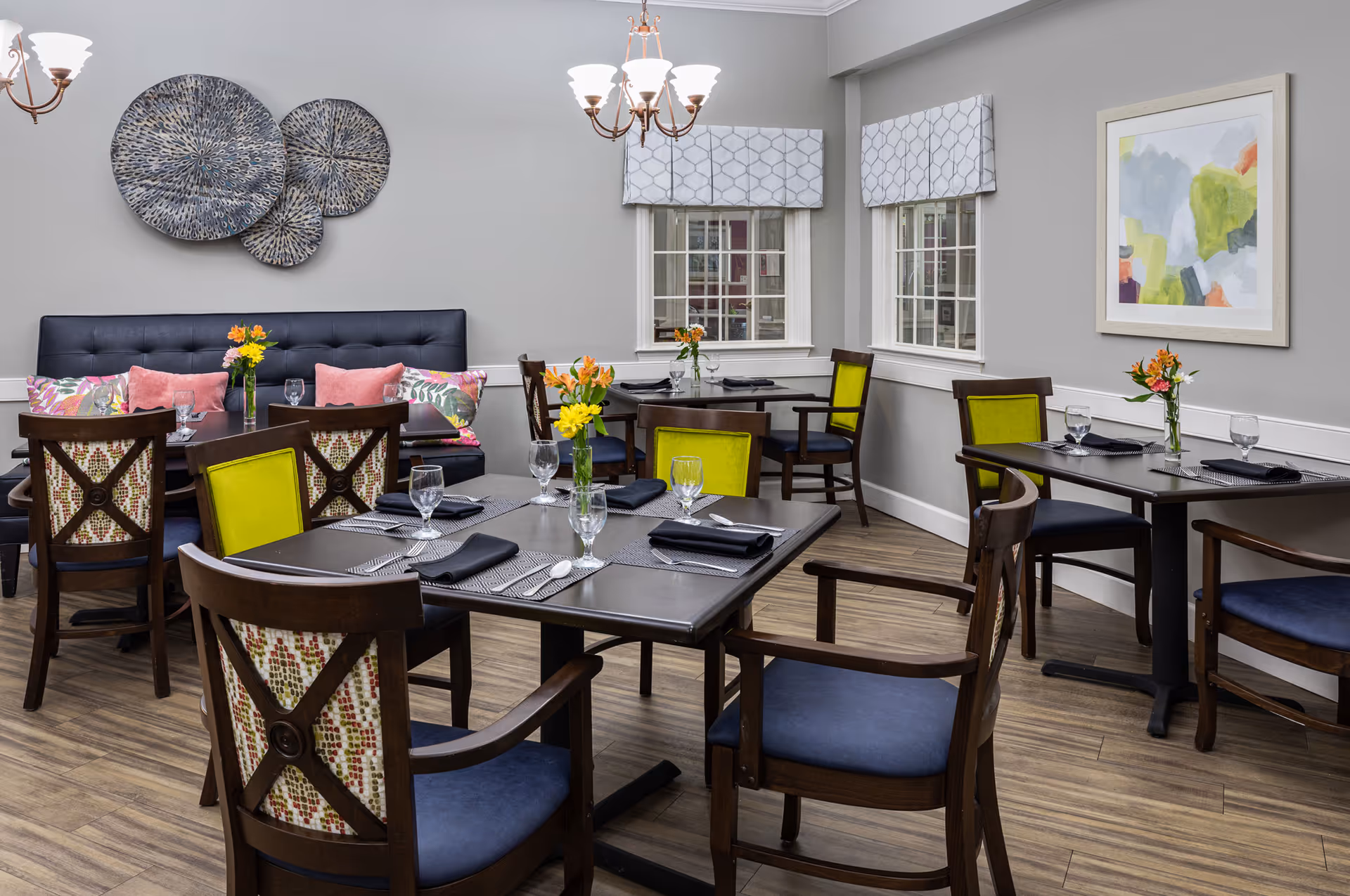 A dining room with several dark wood tables and chairs, some chairs have blue cushions and others have green or patterned backs. The tables are set with placemats, black napkins, silverware, and water glasses. Small vases with colorful flowers are placed on each table. The room has light gray walls, two windows with patterned valances, a framed abstract painting, and decorative circular wall art. Two chandeliers with multiple lights hang from the ceiling.
