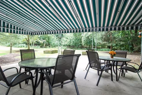 Outdoor patio area with two round glass tables and several black mesh chairs under a green and white striped awning. The patio overlooks a grassy area with trees and shrubs in the background.