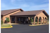 Exterior view of a single-story brick building with a covered entrance supported by arches, a paved driveway, and some landscaping including small bushes and grass.