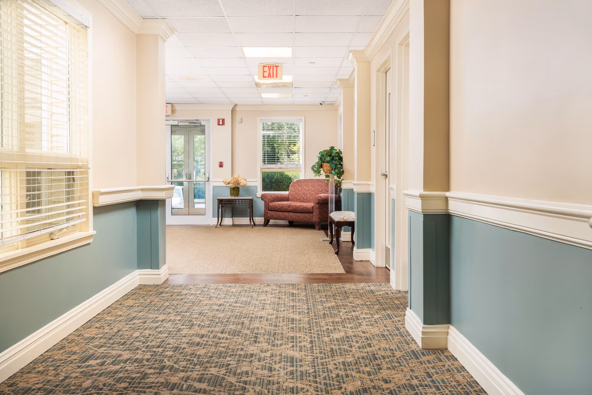 A well-lit hallway in a senior living facility with beige and green walls, patterned carpet, and a seating area at the end featuring a red upholstered loveseat, a small table with a flower arrangement, and a potted plant near a window. There is an exit door with glass panels and an illuminated exit sign above it.