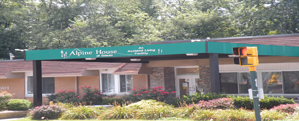 Exterior view of Alpine House of Toledo, an assisted living facility, showing the entrance canopy with green signage and surrounding landscaping with bushes and flowers.