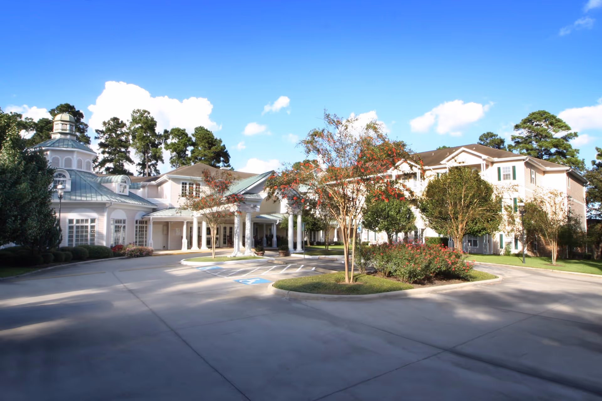 Exterior view of King's Preserve at Kingwood, showing a large building with white walls and green roofs, surrounded by trees and landscaping under a blue sky with some clouds. There is a driveway with parking spaces, including handicapped spots, in front of the building entrance.