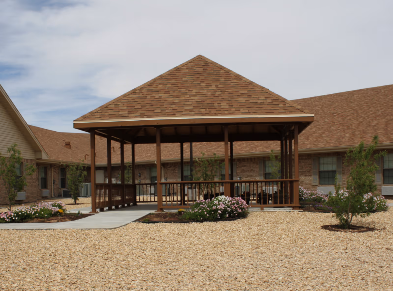A wooden gazebo with a brown shingled roof situated in a landscaped courtyard with gravel ground cover, small bushes, and flowers. Surrounding the gazebo are single-story brick buildings with windows and air conditioning units.